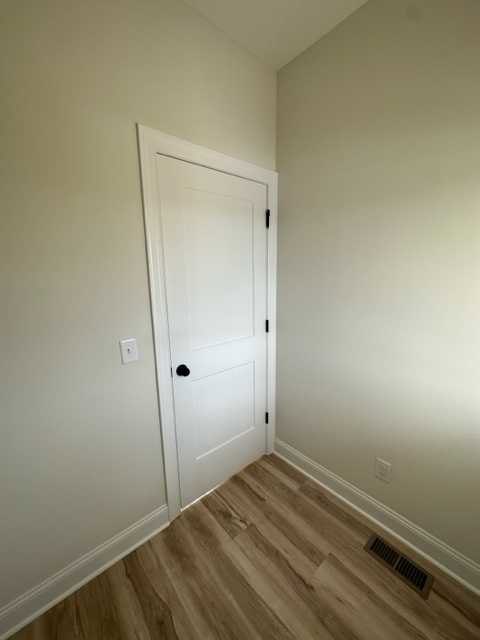 White door with black knob and hinges set in a white wall, wood laminate flooring, wall vent, and light switch visible in corner of room.
