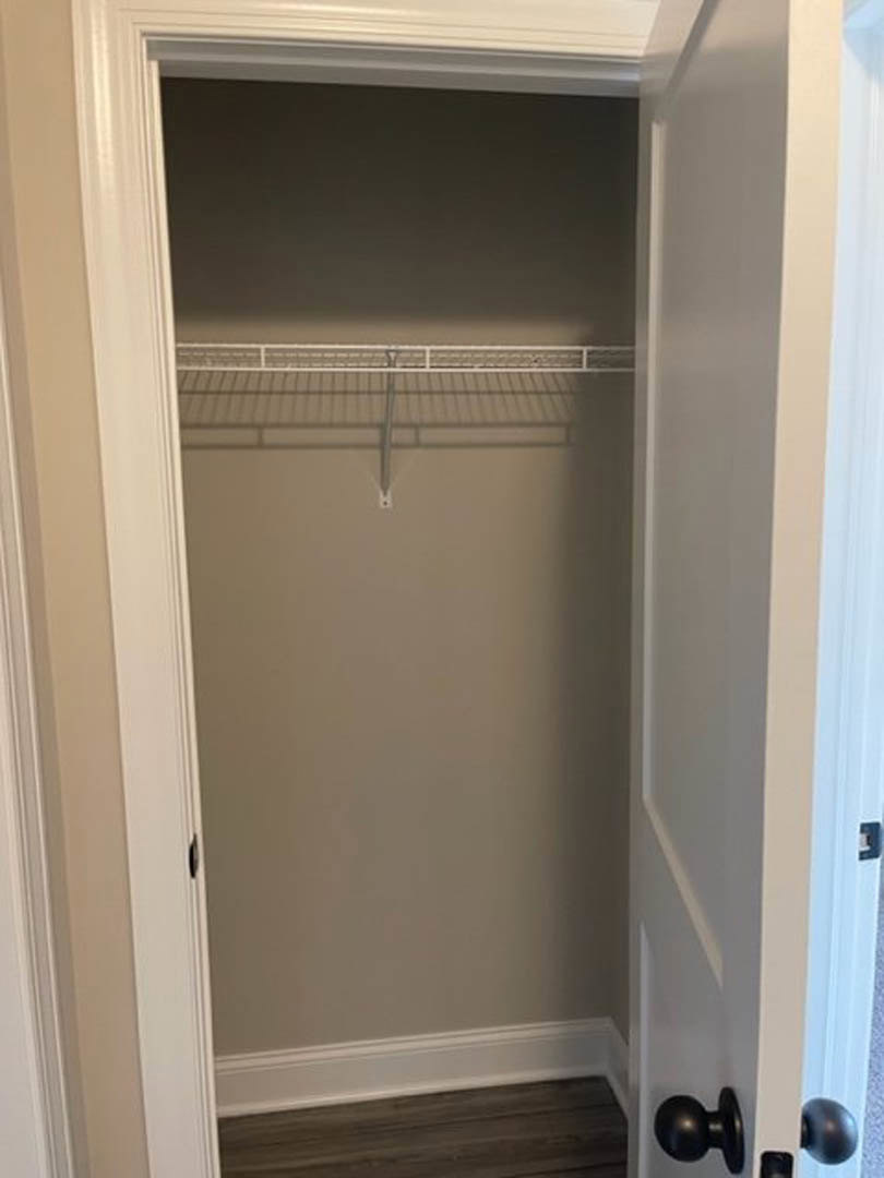 White closet with built-in shelf, black door knob on white door, wood flooring, and wall-mounted light fixture