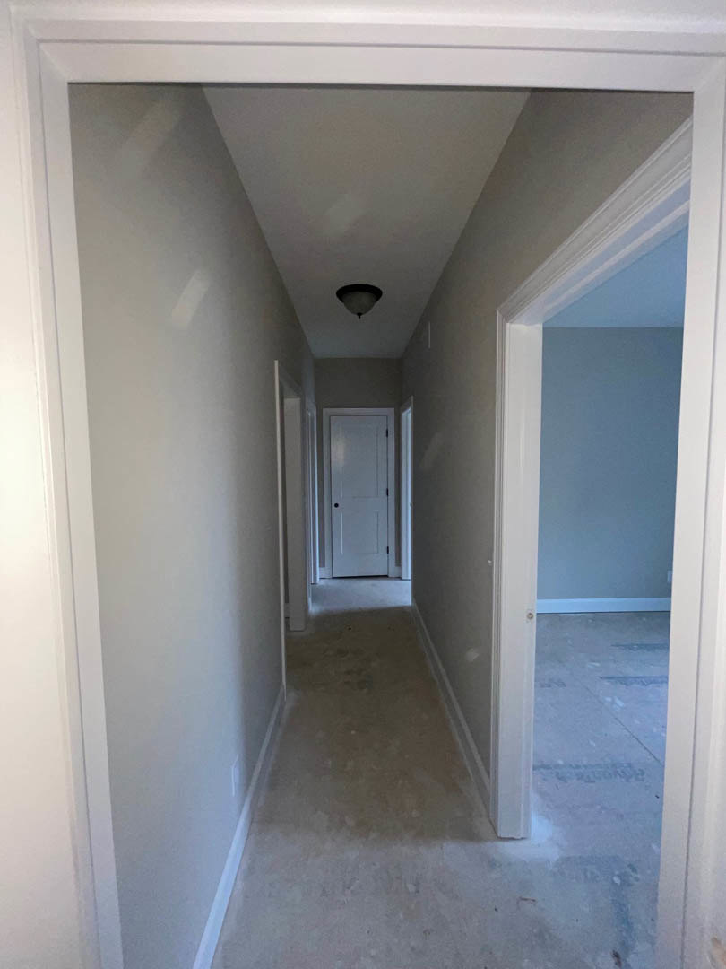 Hallway with smooth white walls, white door featuring a black handle, open doorway leading to a room, ceiling light fixture, natural light streaming in, white baseboards