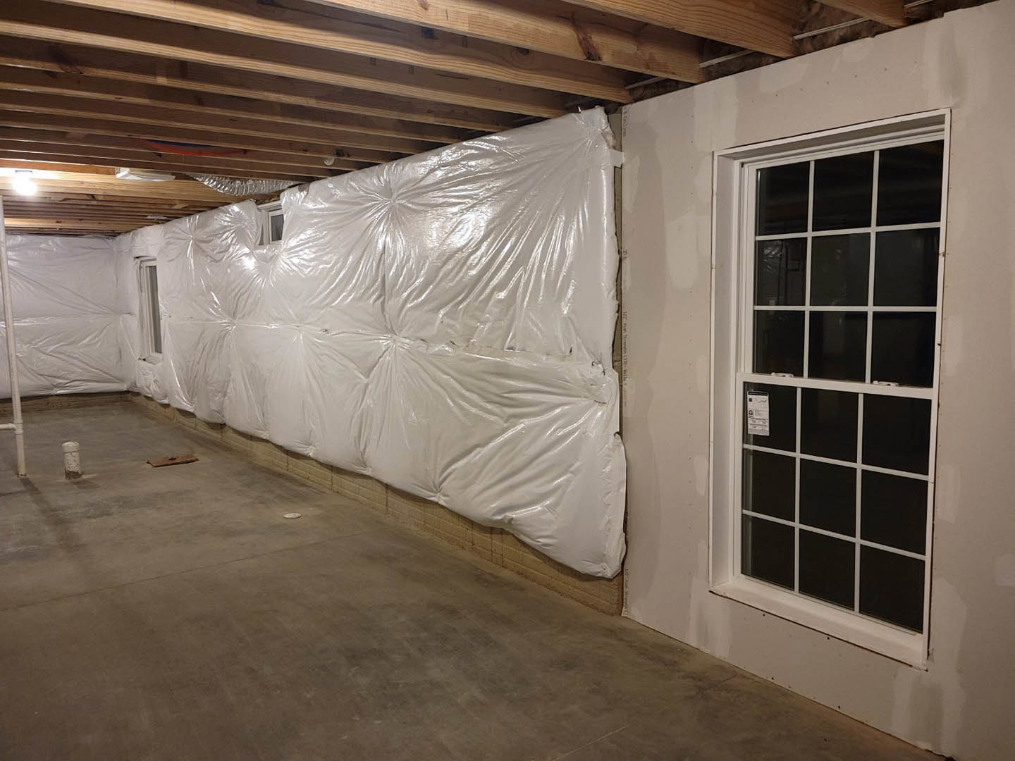 White plastic sheeting covers a wall, white-framed window partially visible, wooden ceiling draped with white fabric, black square screen with white trim on wall, unfinished