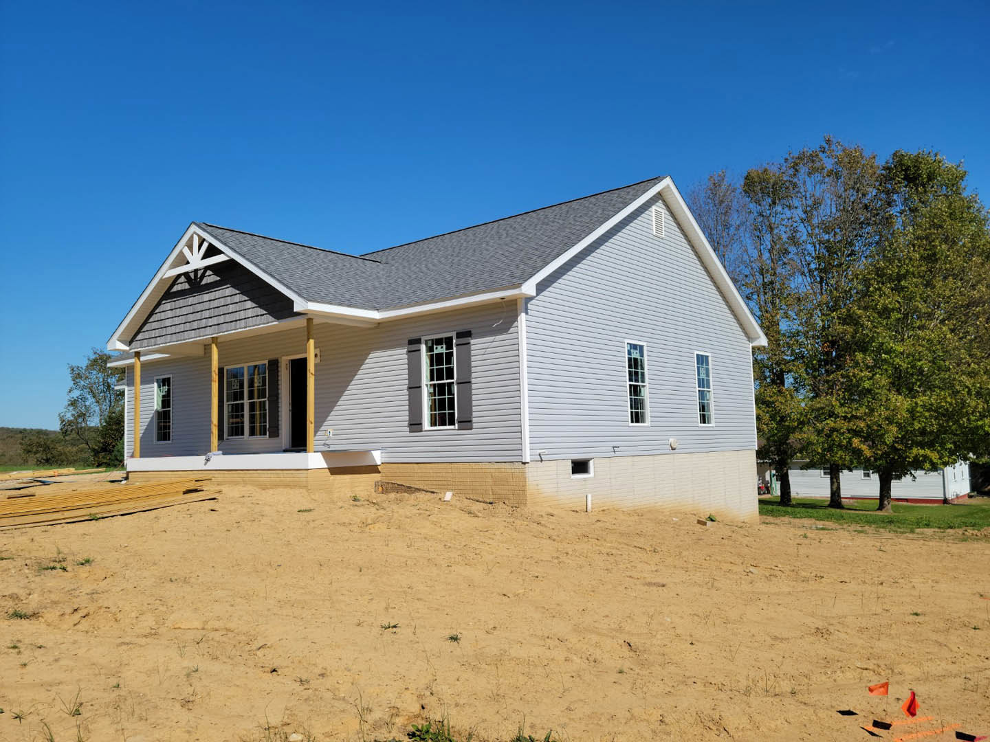 Partially built house with exposed framing and white window frames, surrounded by dirt and construction materials, set against a blue sky with trees in the background