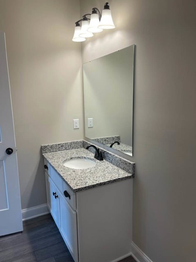 Bathroom with white tile walls, rectangular mirror above a stone countertop sink, chrome faucet, wall-mounted light fixtures, and white cabinetry.