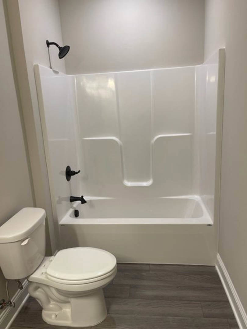 Freestanding white bathtub with matte black faucet, white toilet with closed lid, gray tile floor, white subway tile walls, minimalist bathroom design