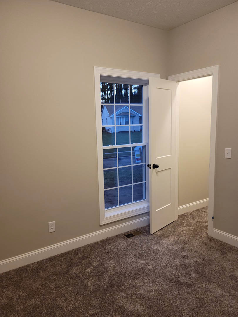 White door with matching frame open into a carpeted room, brown flooring and plaster walls visible, window offers view of neighboring house.