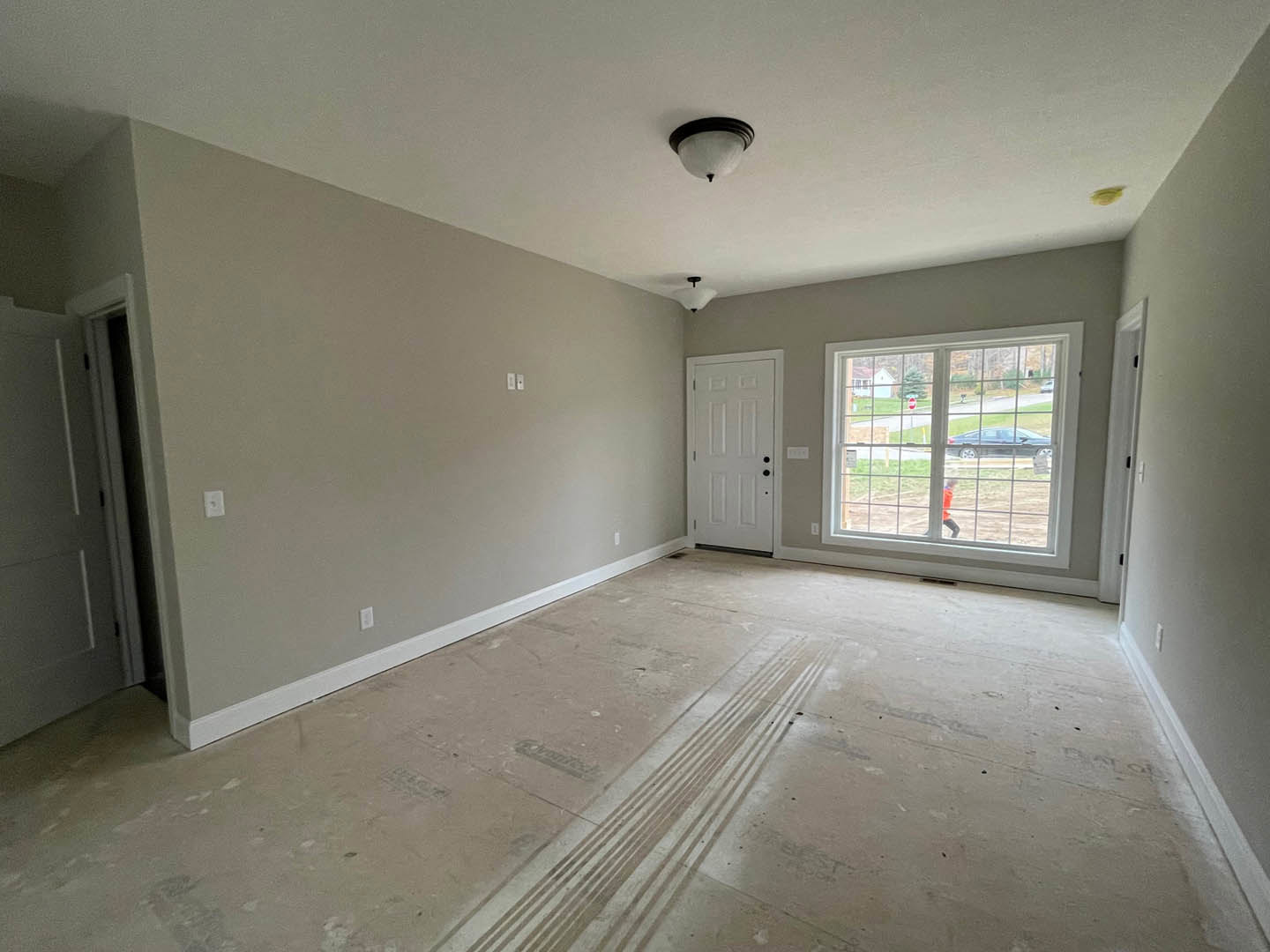 White paneled door with black hardware, large window letting in daylight, light fixture mounted on plaster wall, smooth flooring with visible tire marks, car parked behind metal