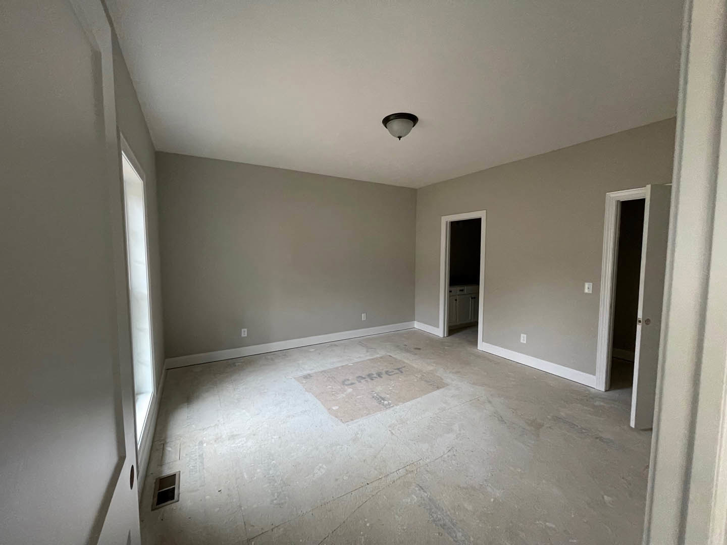 White plaster walls, square window with dark trim, light fixture with white shade, wooden door, patterned rug on hardwood floor