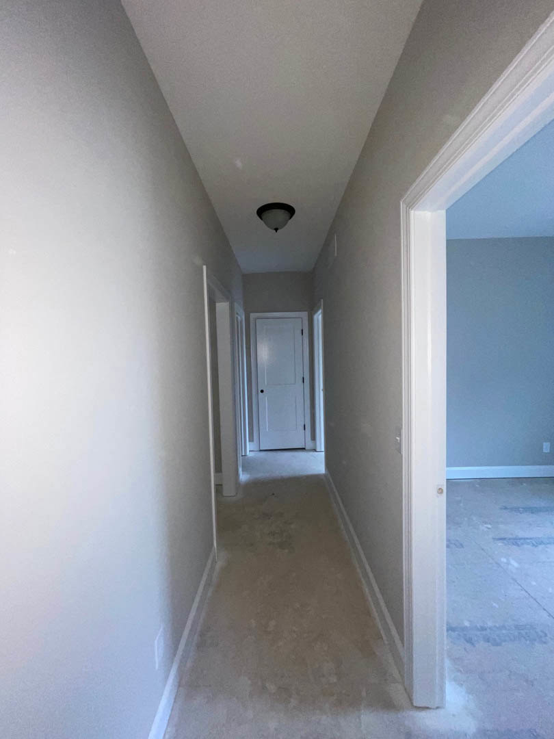 Hallway with white plaster walls, wood flooring, white door with black handle, ceiling light fixture, window showing blue sky and clouds