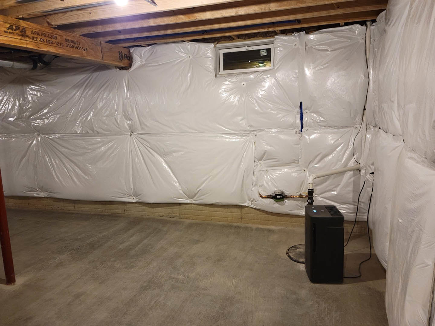 Basement with plastic sheeting covering the walls, exposed wooden beams with printed text, a black rectangular waste container with a handle, window fitted with a black screen, and