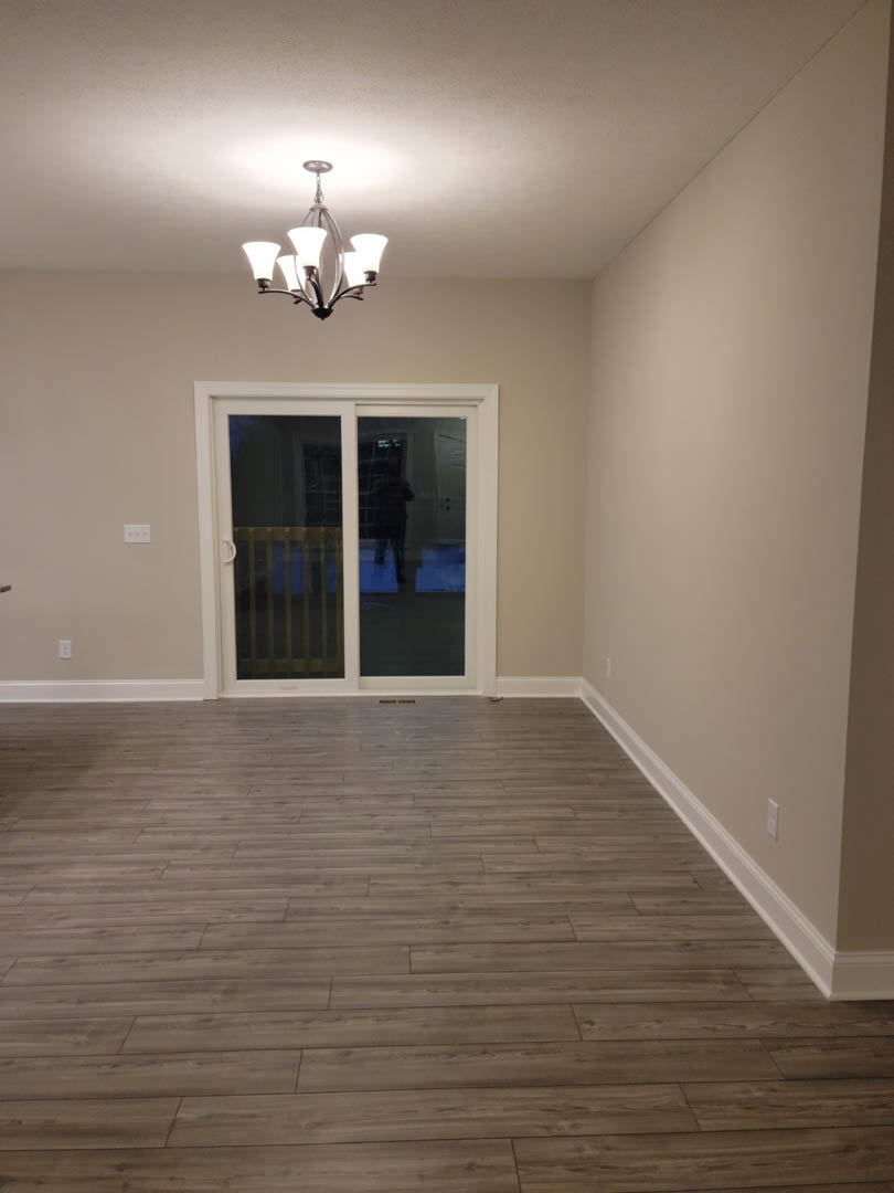 Open living area with wide plank wood flooring, white plaster walls, sliding glass door with white frame, modern chandelier overhead, large window, blurred figure walking past