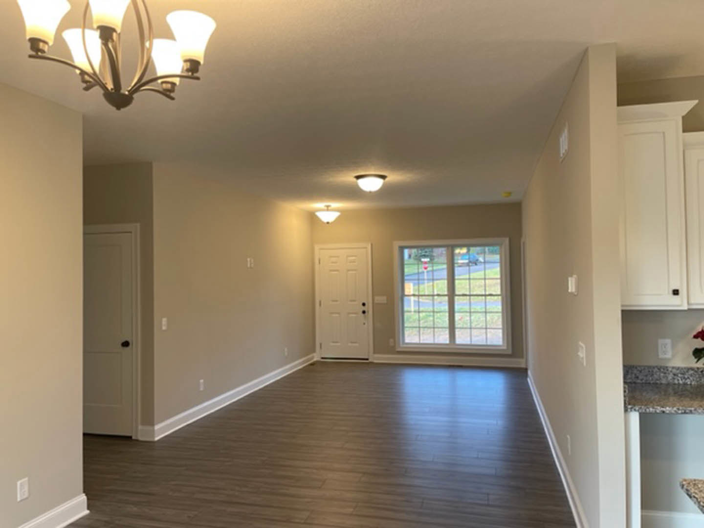 Hallway with light wood flooring, white walls, white door with black hardware, window overlooking street and lawn, modern chandelier overhead