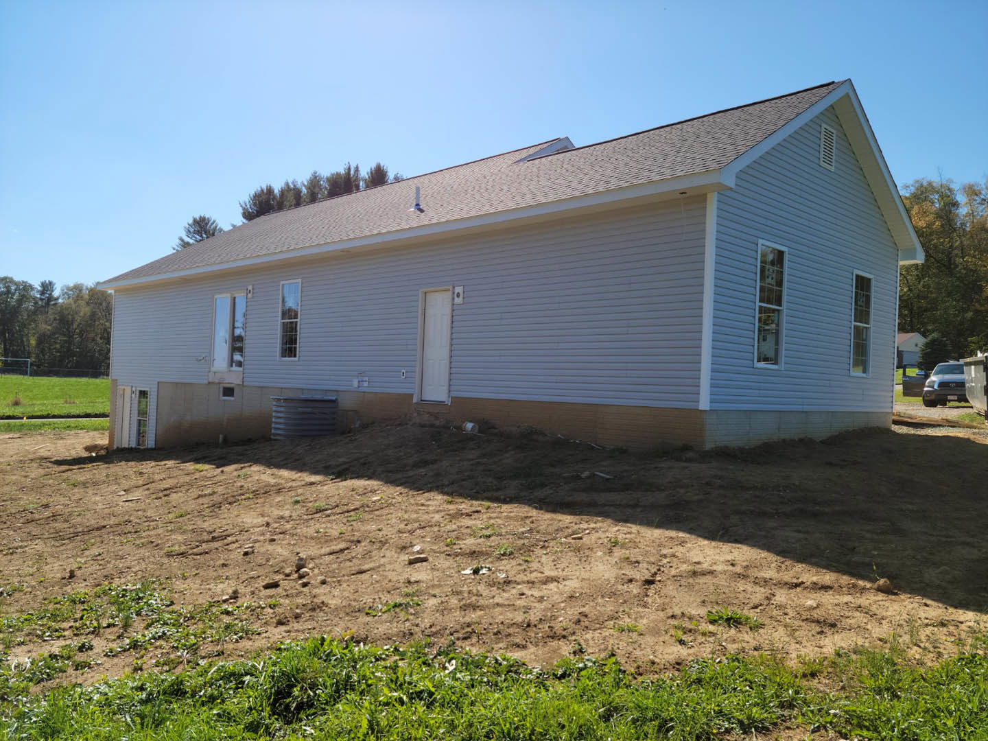 Two-story house with white-framed windows and door, situated on a dirt hill with patches of grass, brick retaining wall, and a parked car in the foreground.