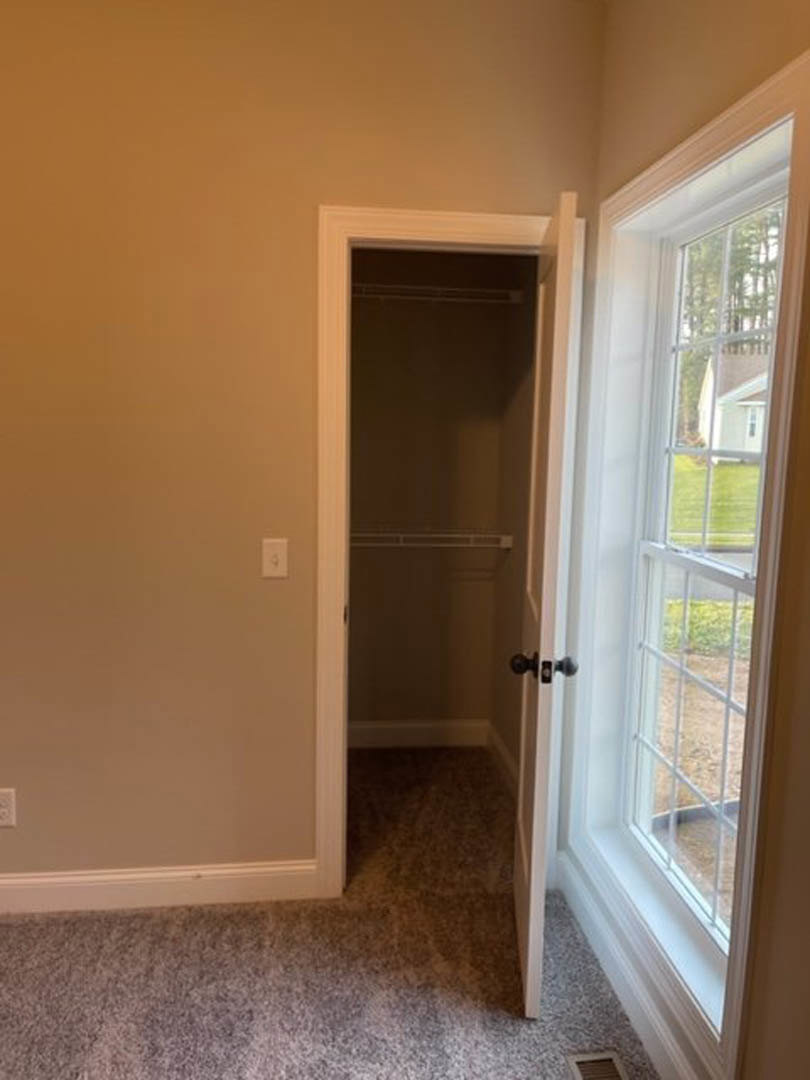 Carpeted bedroom with white closet door, built-in shelf, large window overlooking grassy yard, light switch on wall, sunlight reflecting on window glass