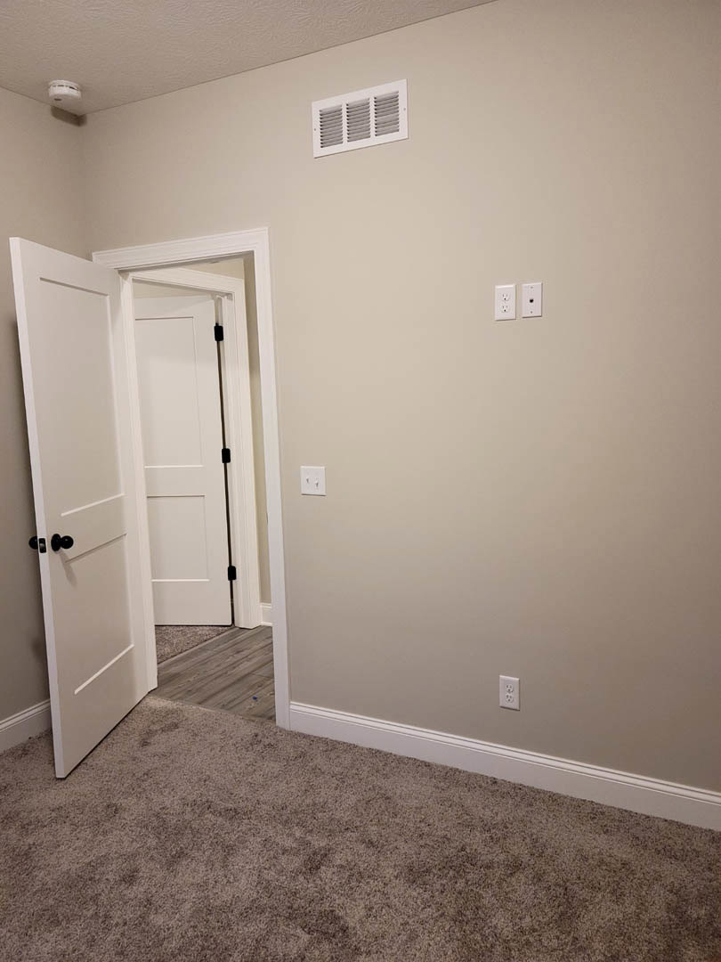 White paneled door open to a carpeted room with light-colored walls, wall vent, and simple door handle