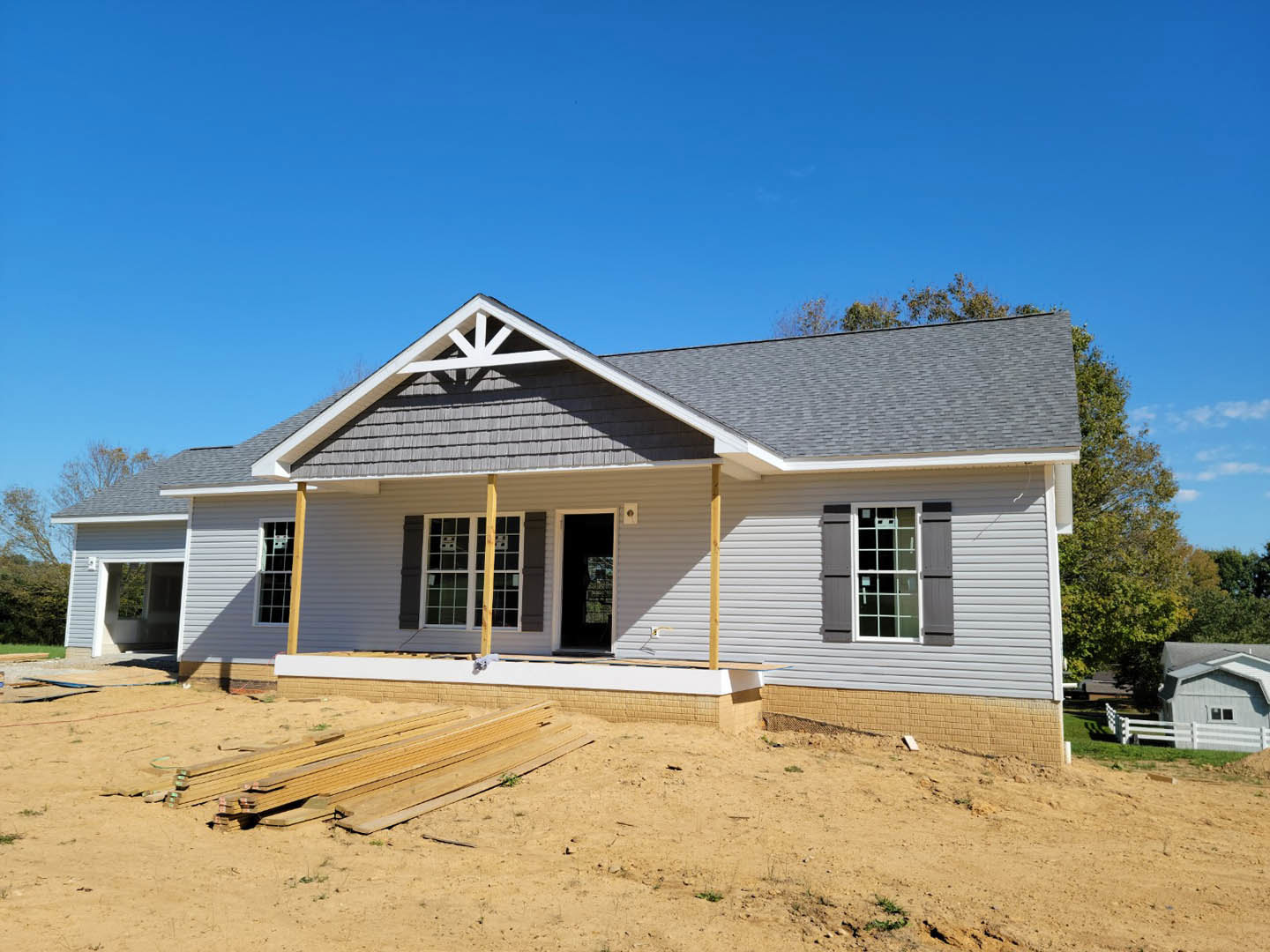 Partially built house with exposed framing, roof overhang, dirt yard, wood planks stacked nearby, person standing in doorway, white siding, and large windows.