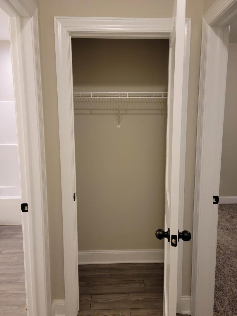 Closet with white walls, wood floor and white trim, built-in shelf, white door with silver handle, carpet visible in adjacent room