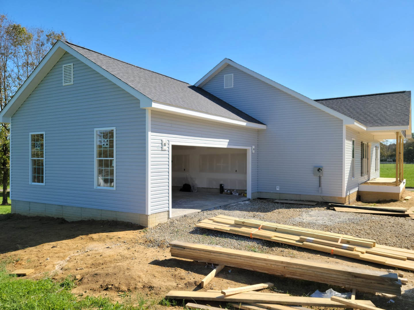 Partially built house with white-framed windows, attached garage with closed door, unfinished roof, pile of lumber on dirt driveway, blue sky overhead