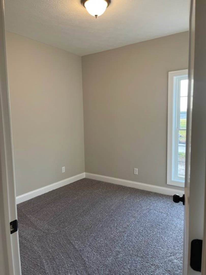 Carpeted room with white trim, open white door, ceiling light fixture, window overlooking grassy yard, black rectangular vent with white border on wall