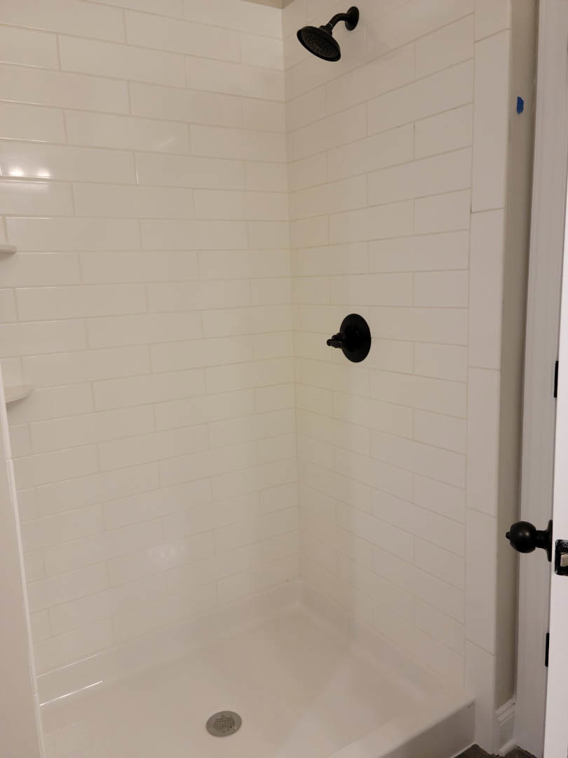 White tiled shower with black shower head and handle, adjacent white bathtub with chrome drain, white door featuring black lever handle