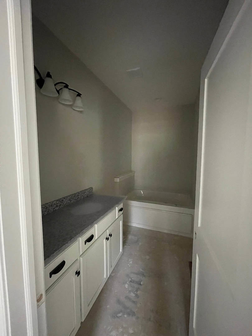 White bathroom with freestanding tub, rectangular sink on stone countertop, black drawer handle, wall-mounted light fixture, white cabinetry, tile floor, and white plaster walls