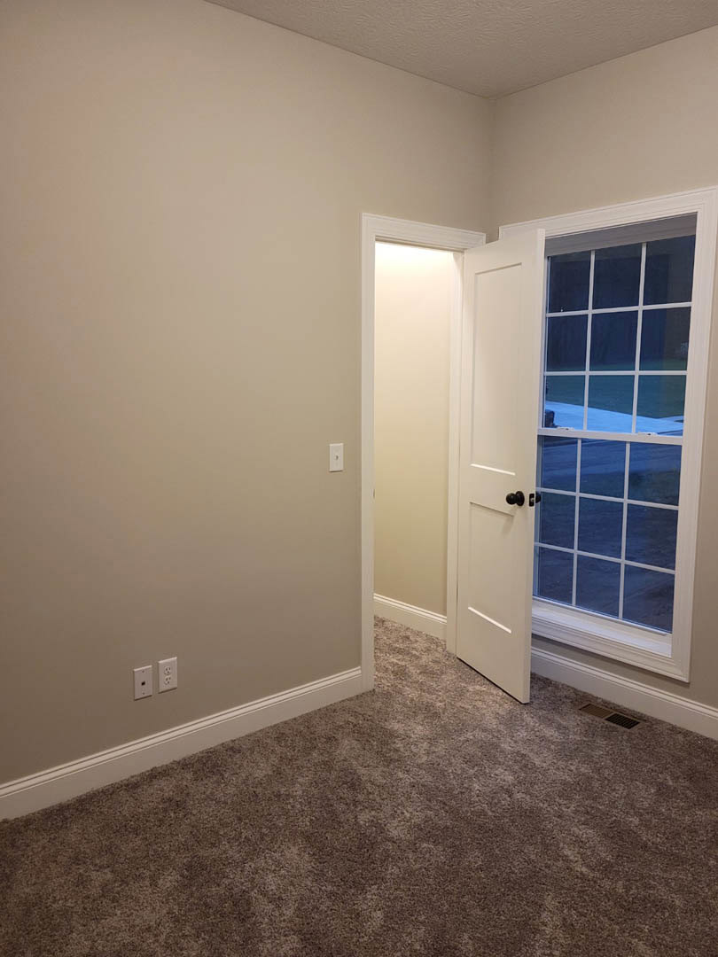 White paneled door with black knob open onto carpeted floor, white door frame and border, wall outlet visible on light-colored plaster wall