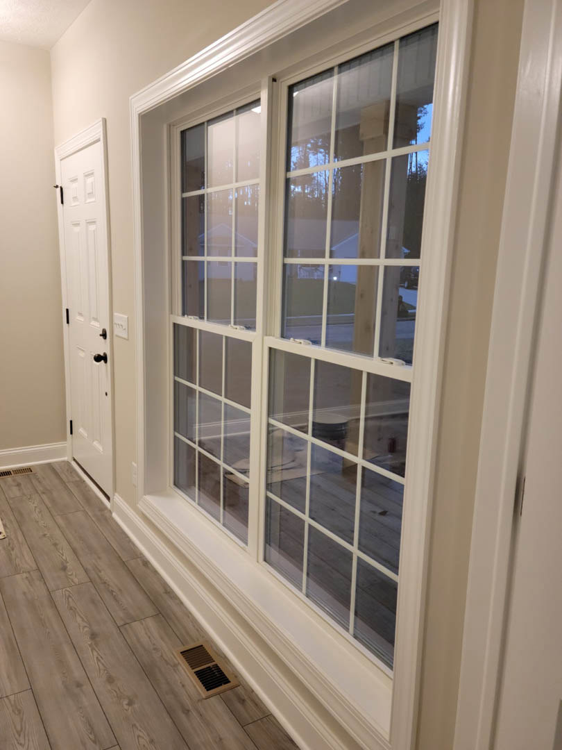 White-trimmed window with closed blinds, wood floor featuring a metal vent, white paneled door with black handles, light-colored walls