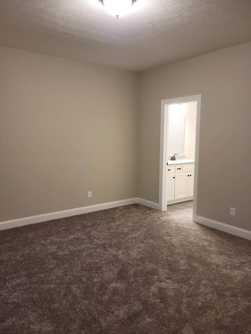 Carpeted room with white walls, open door leading to bathroom with white cabinets and sink, ceiling light fixture, grey carpet, light reflecting on white wall
