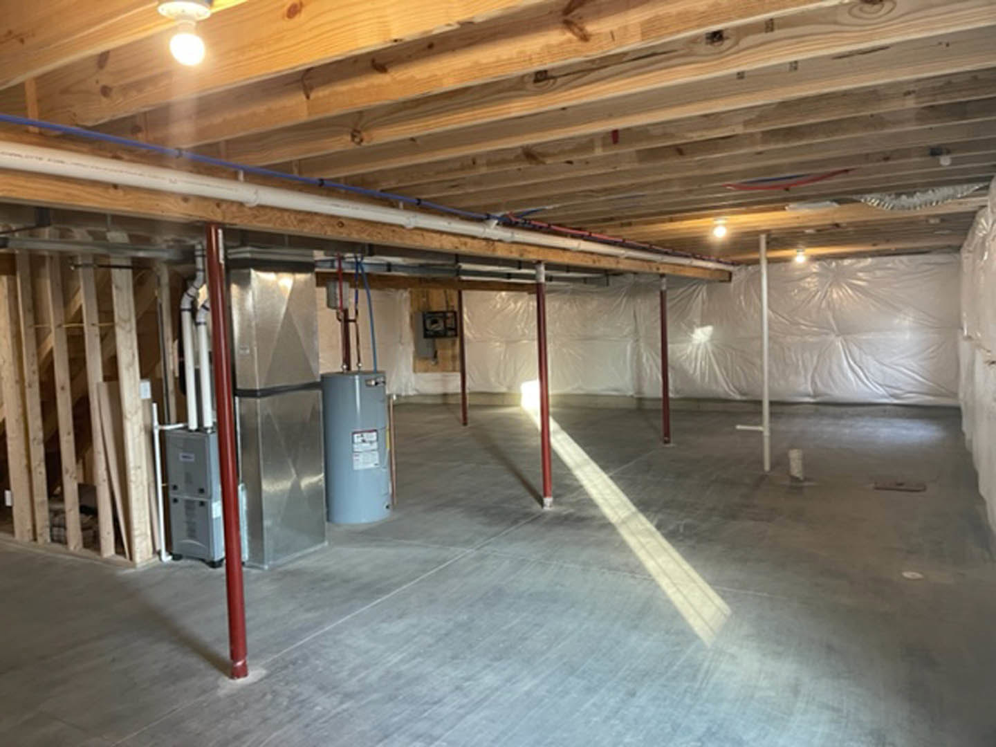 Wood plank ceiling above concrete floor covered with white plastic sheeting, exposed steel beam and unfinished walls in basement interior.