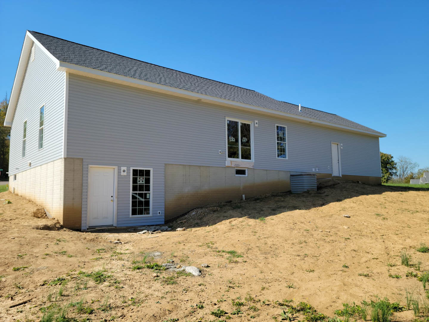 Partially built house with white siding, attached garage, white door featuring yellow handle, dirt foreground, windows reflecting trees, construction sign visible in window, under