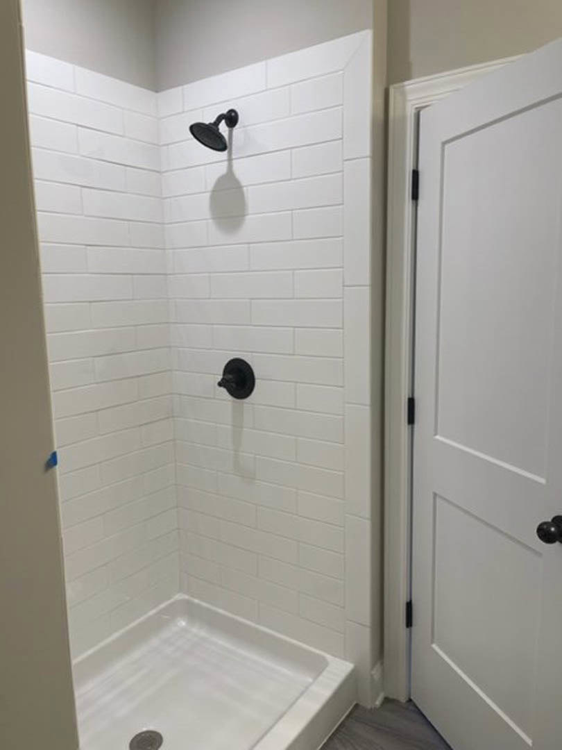 Black shower head mounted on white tile wall above white bathtub, close-up of white door and black door handle visible.
