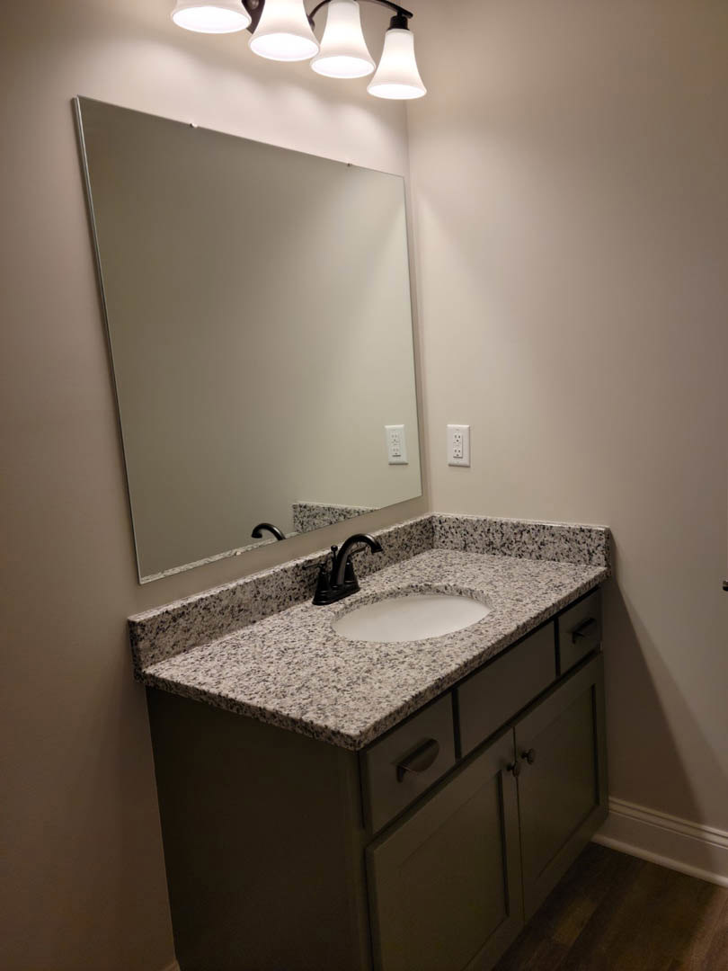 White porcelain sink with chrome faucet set in a stone countertop, rectangular wall mirror above, light fixture reflected in mirror, tile backsplash, and wood cabinetry with