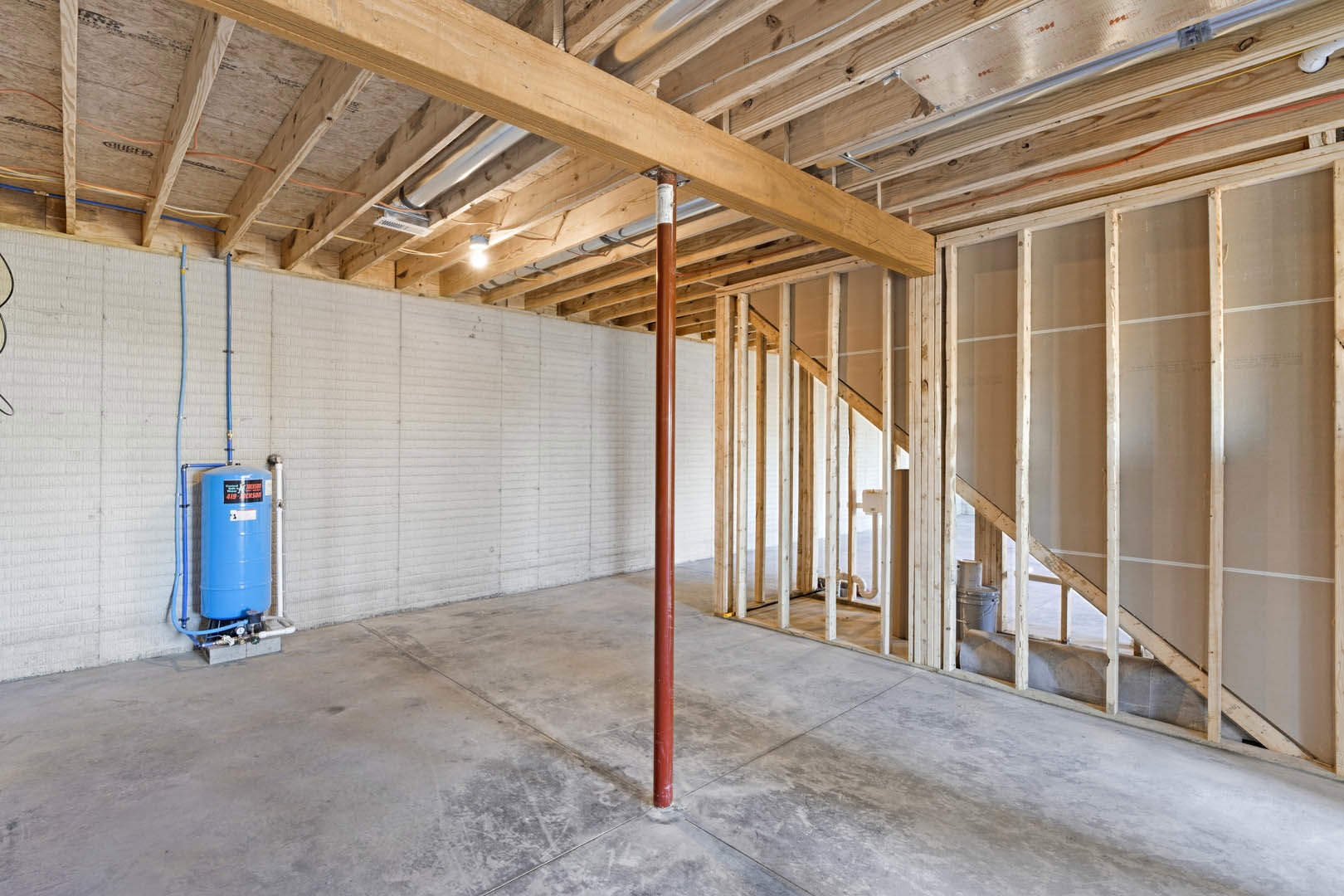 Basement utility room with a large blue water tank, blue metal pole, exposed wooden beam, concrete floor, and unfinished walls.
