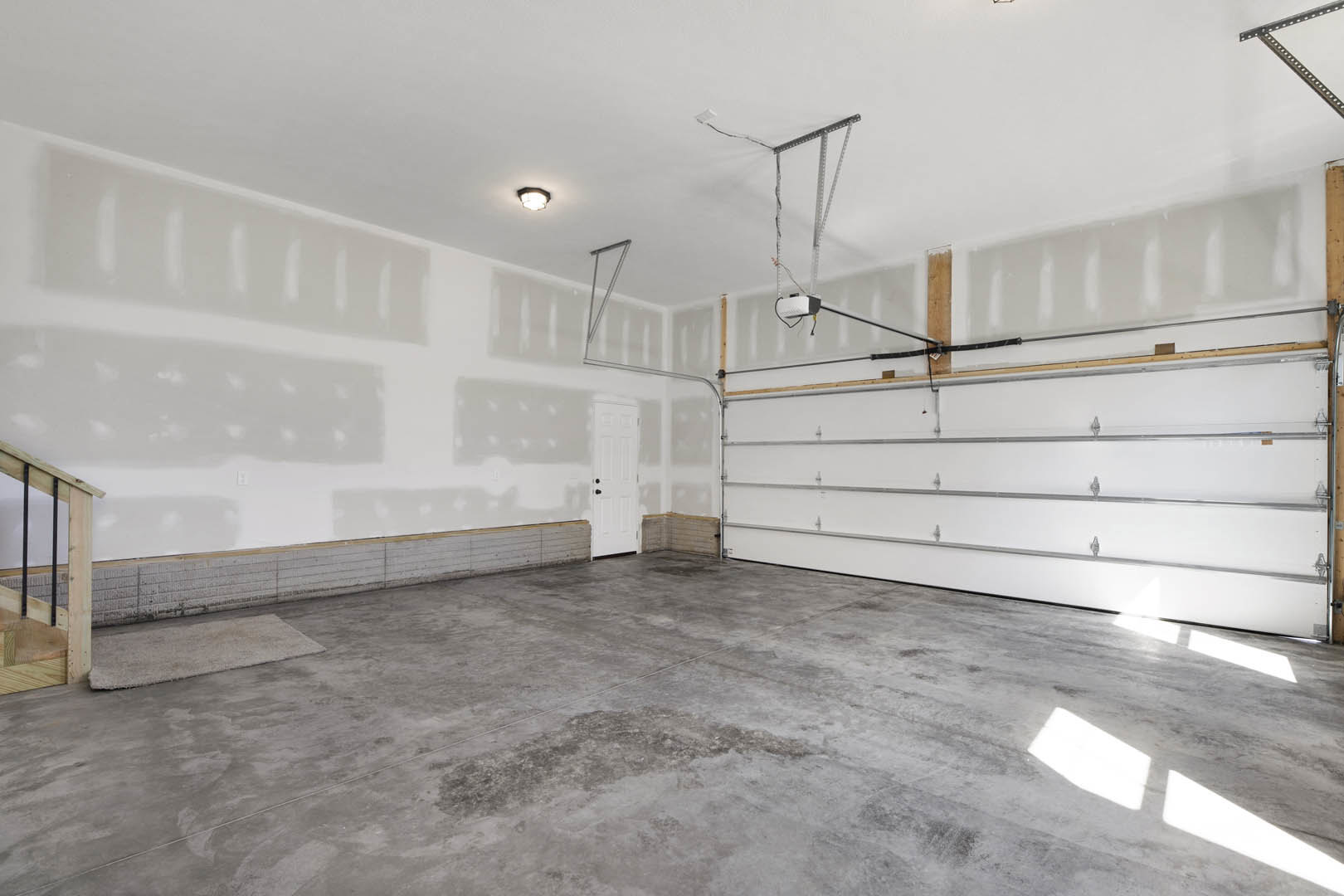 Garage interior with white plaster walls, white door featuring black knobs, concrete floor, overhead lighting, and close-up view of wooden railing