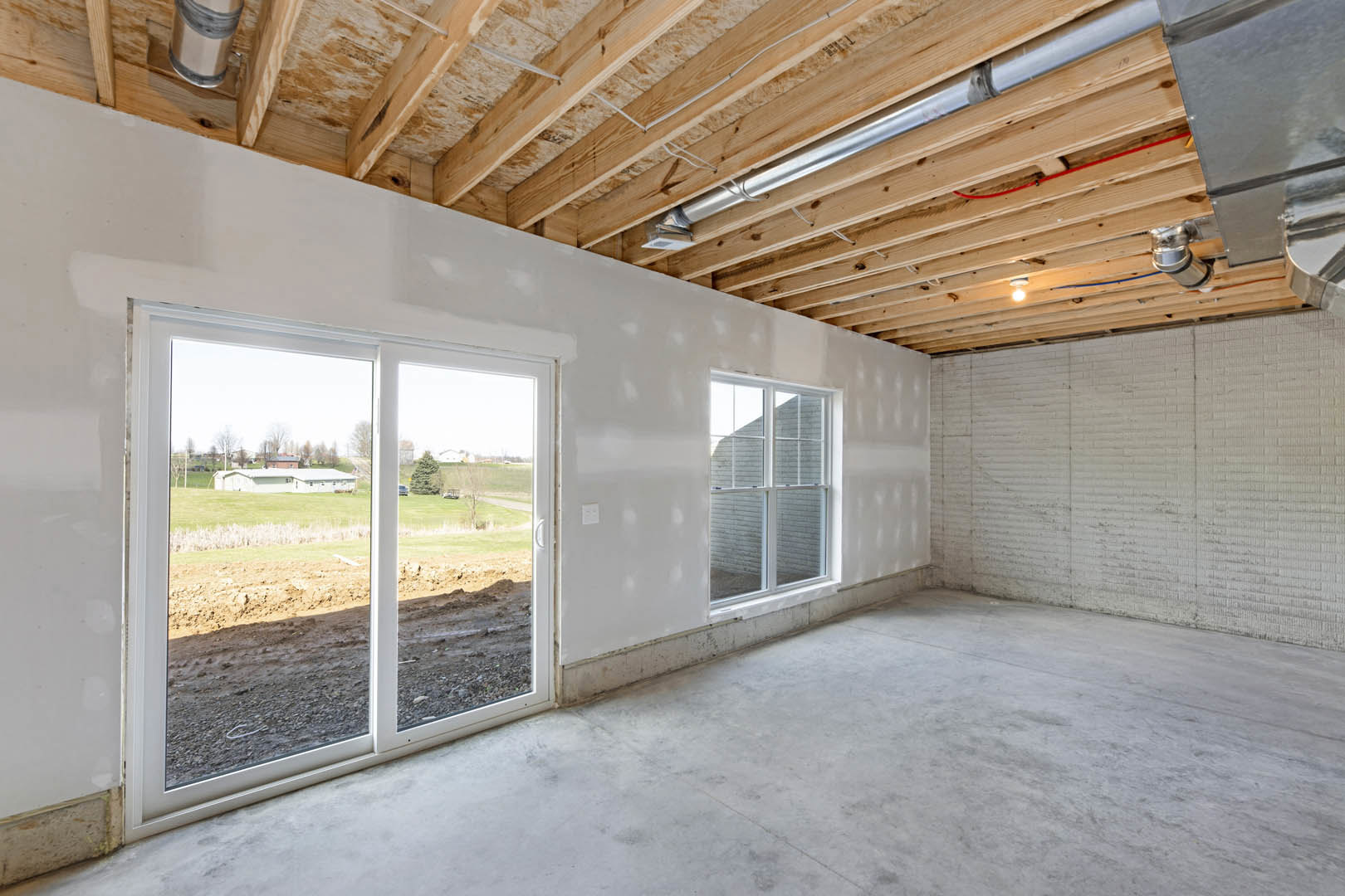 Open room with concrete floor, white brick wall, exposed wood ceiling beams, sliding glass door overlooking grassy field, ceiling pipe visible.
