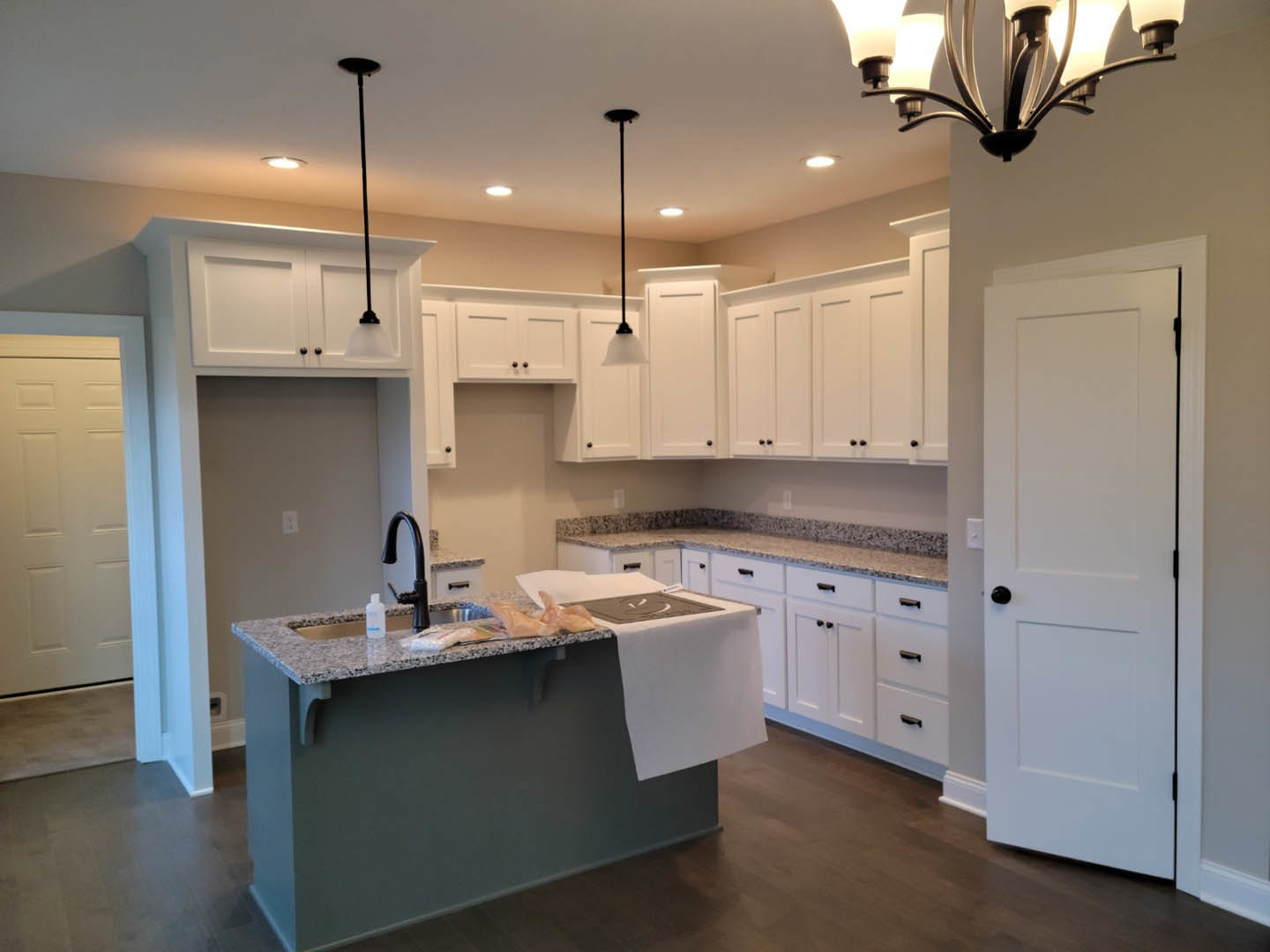White kitchen cabinets, stainless steel sink set in a light stone countertop, towel draped beside the basin, wood flooring, white paneled door, modern ceiling lamp.