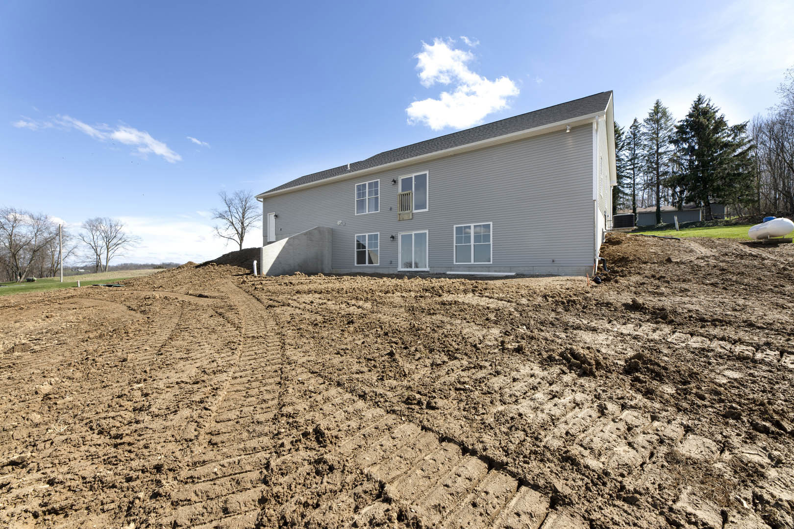 Grey house exterior with white-framed windows, dirt hill and tire tracks in muddy ground, white tank with blue top, scattered trees, blue sky with clouds