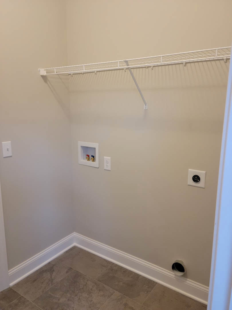 White tile floor and plaster walls with circular vent, black outlet, brass faucets, and white wall-mounted shelf in a bathroom.