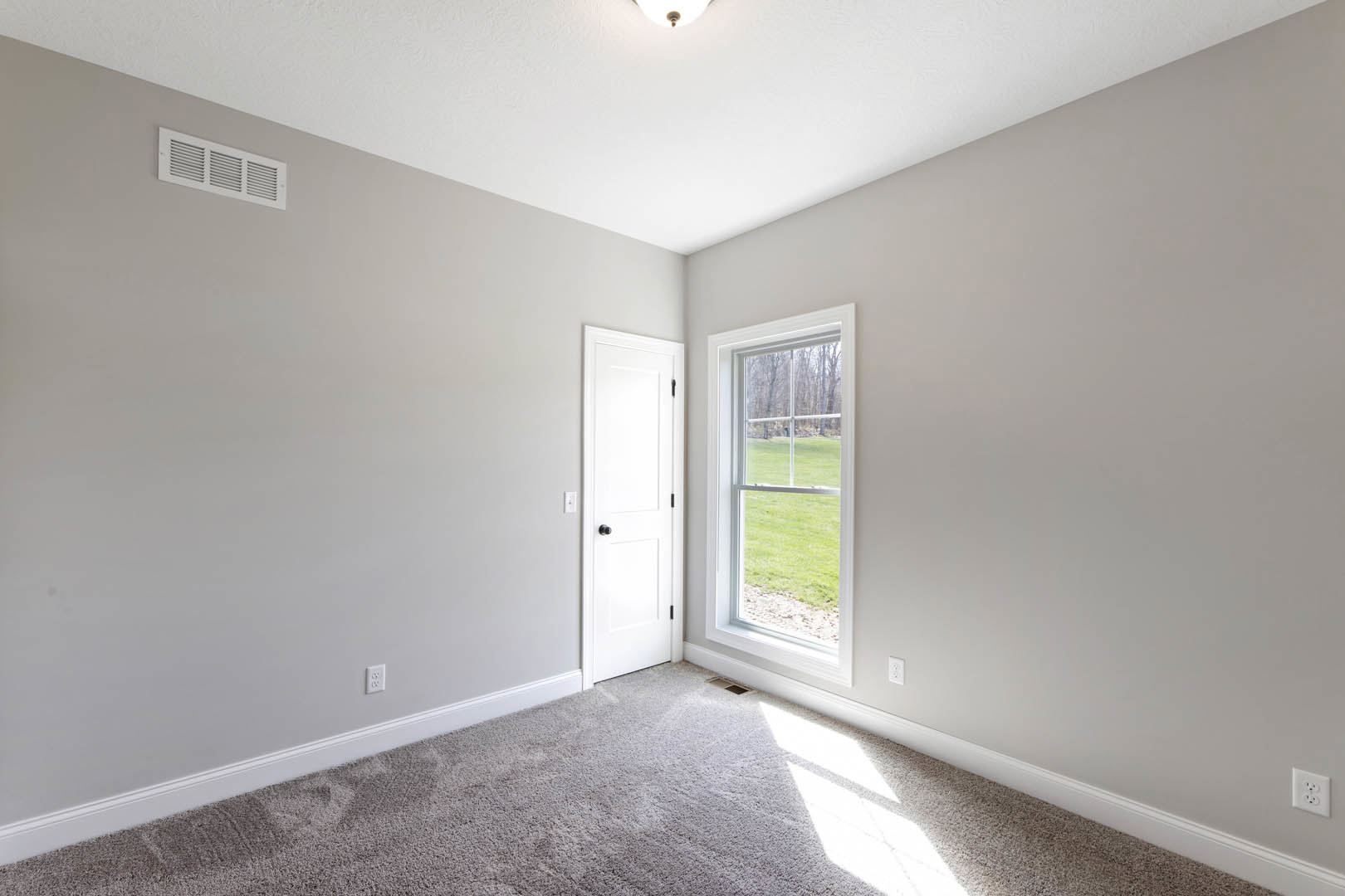 Carpeted floor in a neutral-toned room featuring a white door with black knob, window overlooking grassy yard, white electrical outlet, and wall vent