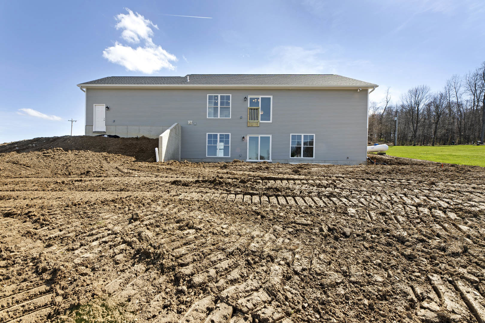 Grey house with white trim, white-framed windows, muddy ground with patches of grass, blue sky and clouds overhead, trees in background