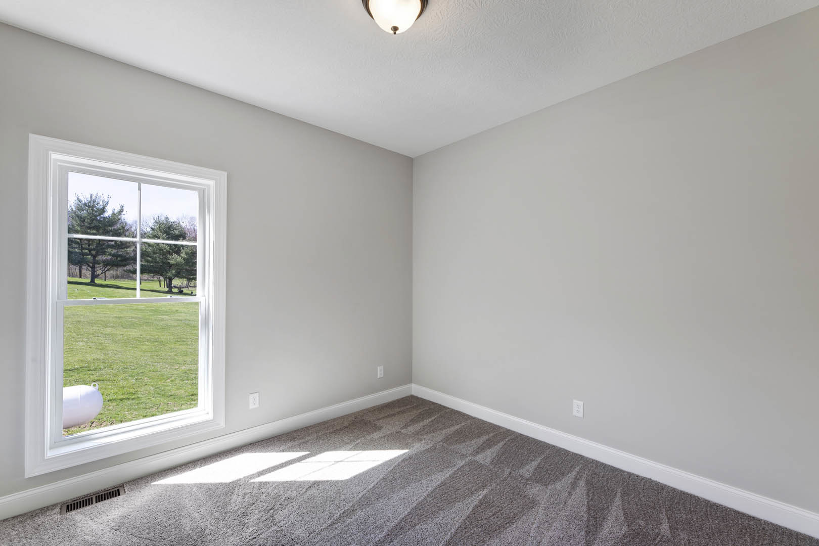 Sunlit room with carpeted floor, large window overlooking green lawn, white walls with crown molding, ceiling fan, and minimal window coverings.