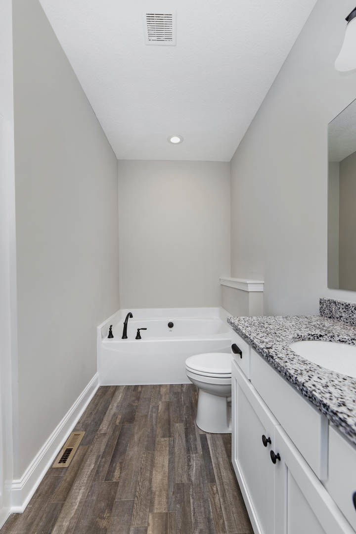 Bathroom featuring a marble countertop with undermount sink, wood flooring with floor vent, white bathtub with black faucet, and close-up view of a toilet.