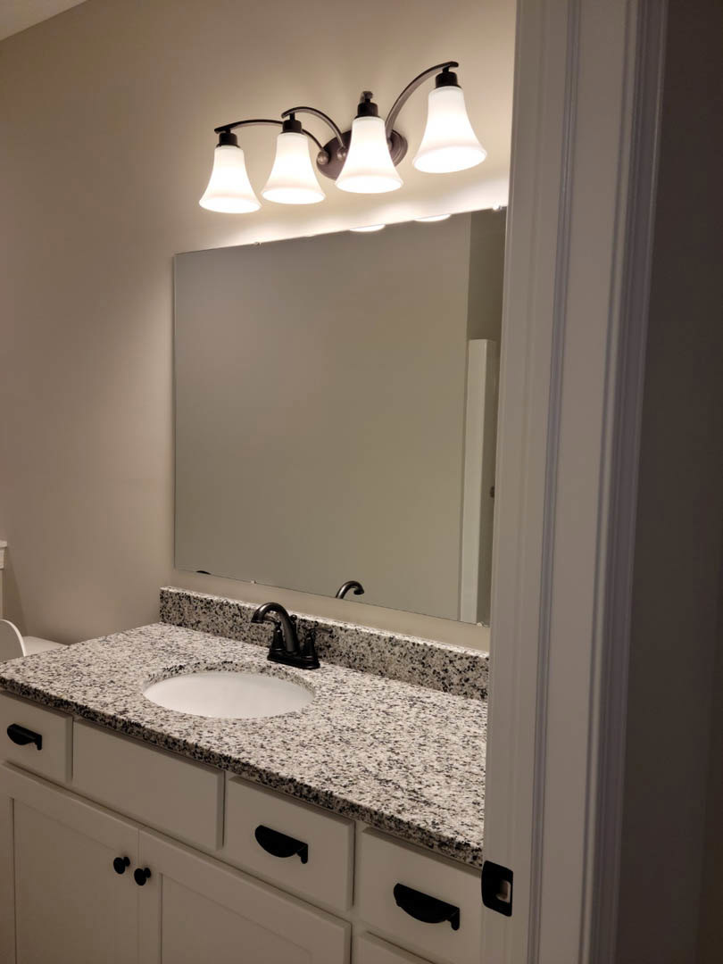 Marble countertop with white sink and black speckled surface, black faucet, large mirror above, light fixture mounted on wall, white board visible in background, tile backsplash