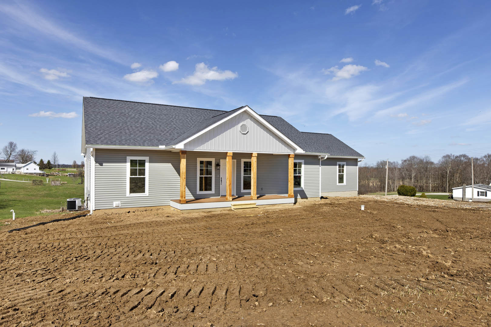 Two-story house with covered porch, white-framed windows, and wooden support poles, set beside a dirt field with tire tracks; white and black construction equipment parked nearby