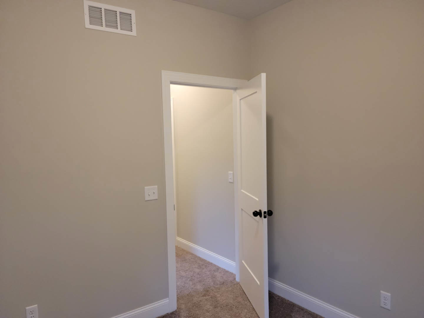 White paneled door with black handle partially open in a room with light-colored plaster walls, visible light switch, electrical outlet, and wall vent; hardwood flooring and white