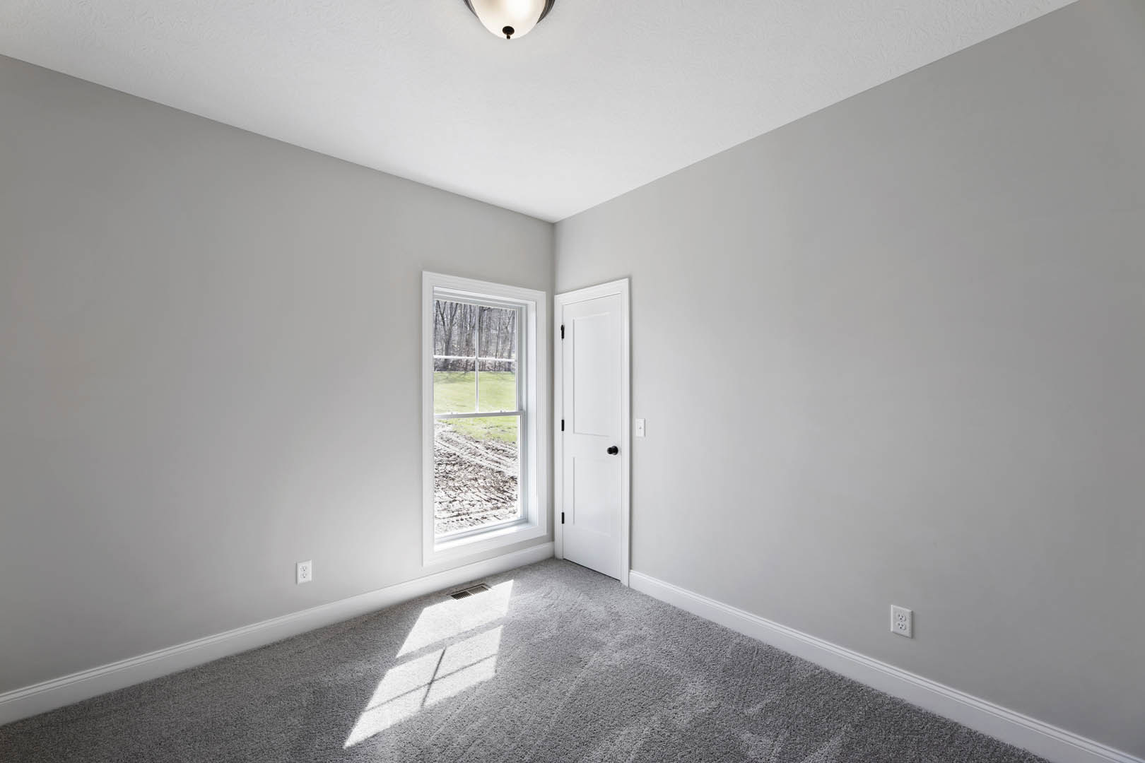 Grey carpeted bedroom with white walls, white door featuring black hardware, ceiling light fixture, and window overlooking a grassy field and trees