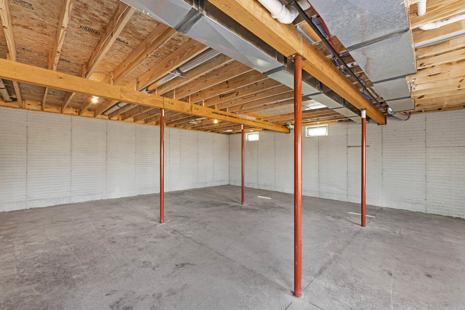 Open room with exposed red metal poles, white painted wall, concrete floor, ceiling featuring visible metal pipes, and window allowing natural light.