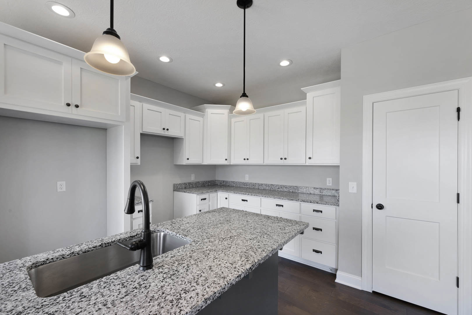 White kitchen cabinets with granite countertops, stainless steel sink and black faucet, tile backsplash, white door with black knob, pendant light fixture, and modern appliances.