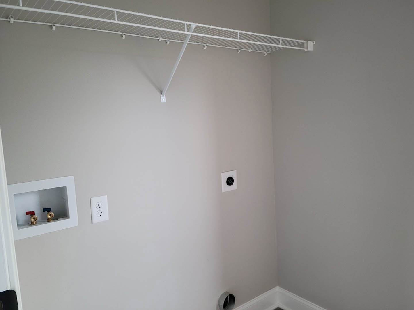 White plaster bathroom wall with built-in white shelf, brass faucets, white window, electrical outlet, black circular fixture, and exposed grey pipe.