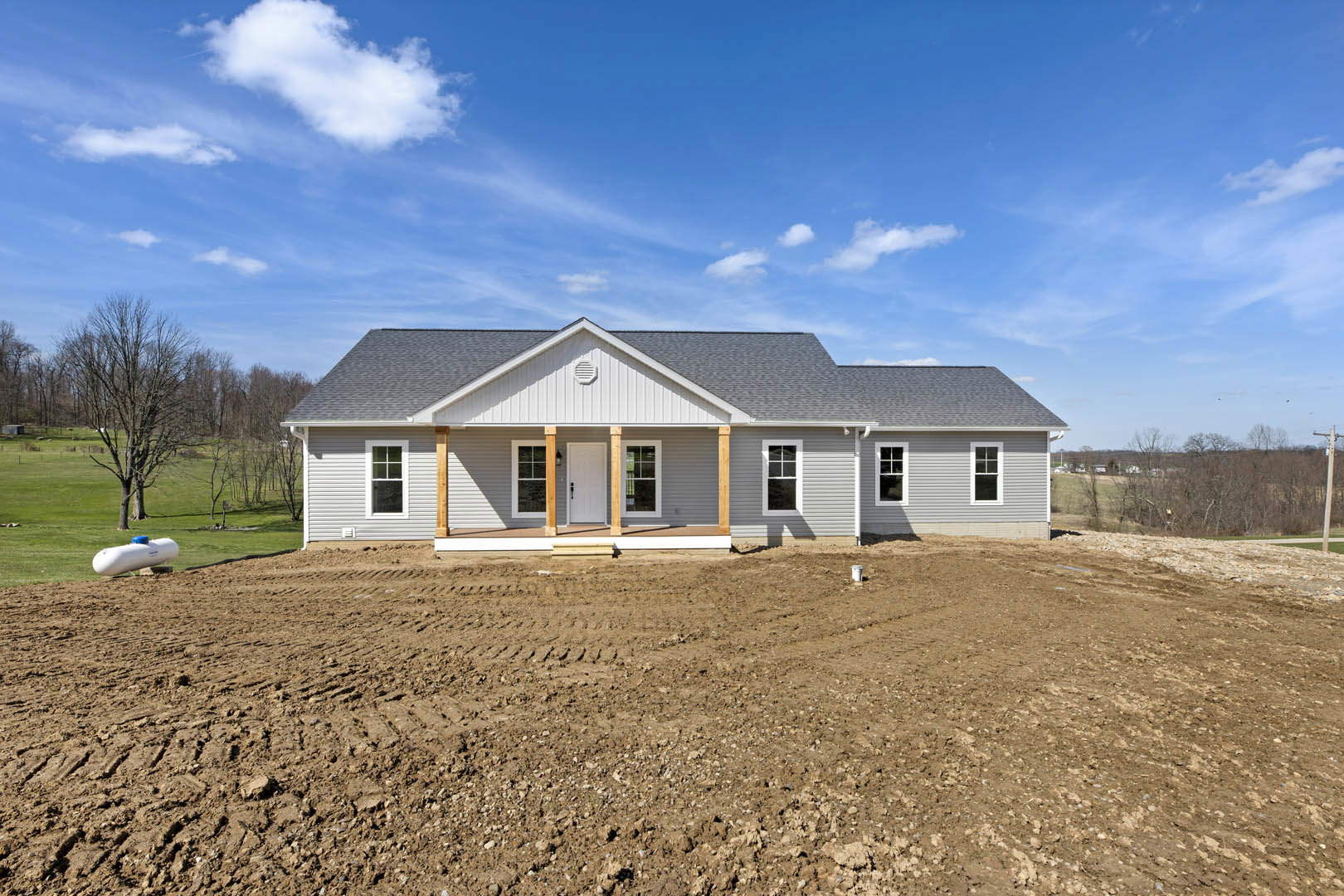 Single-story house with white doors, surrounded by a dirt field, leafless tree, white pole, and white tank with blue cap under a cloudy sky