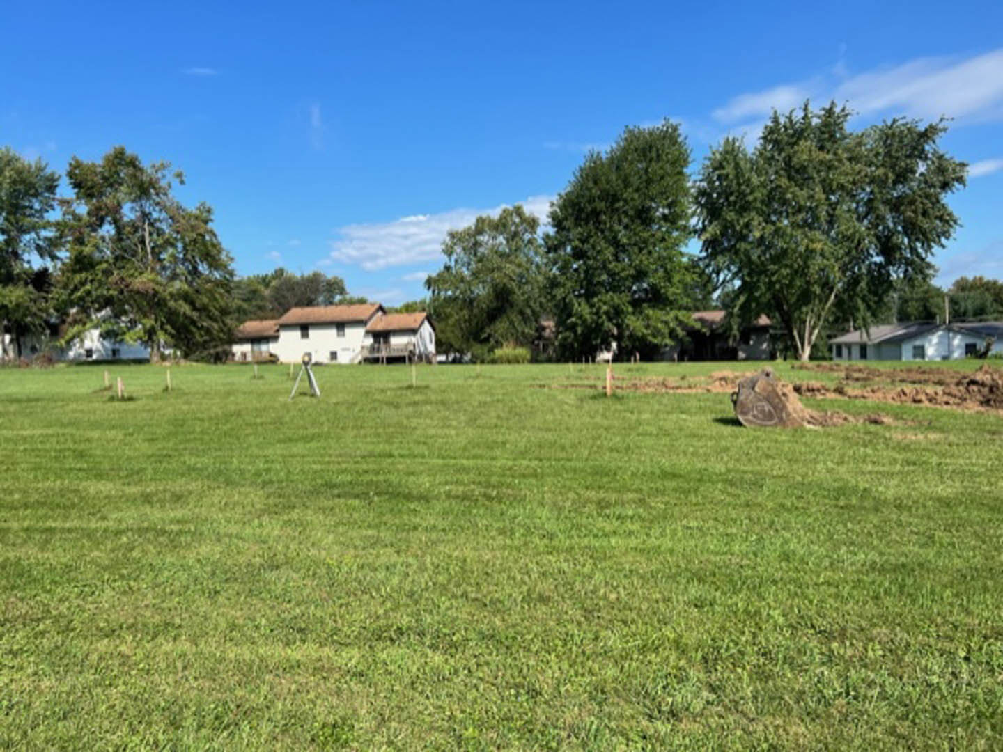 Expansive green lawn bordered by mature trees, distant houses with white siding and red roofs under a blue sky with scattered clouds