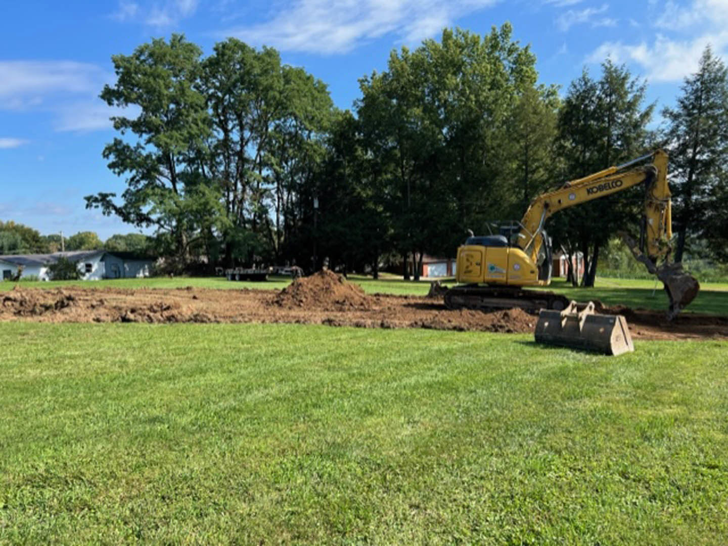 Yellow excavator with large arm parked on grassy field, surrounded by trees and cloudy sky in rural area
